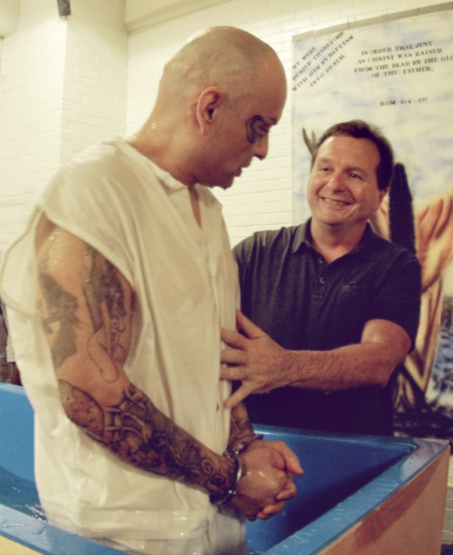 A tattooed man being baptized inside a prison unit, the man baptizing him smiling beside him