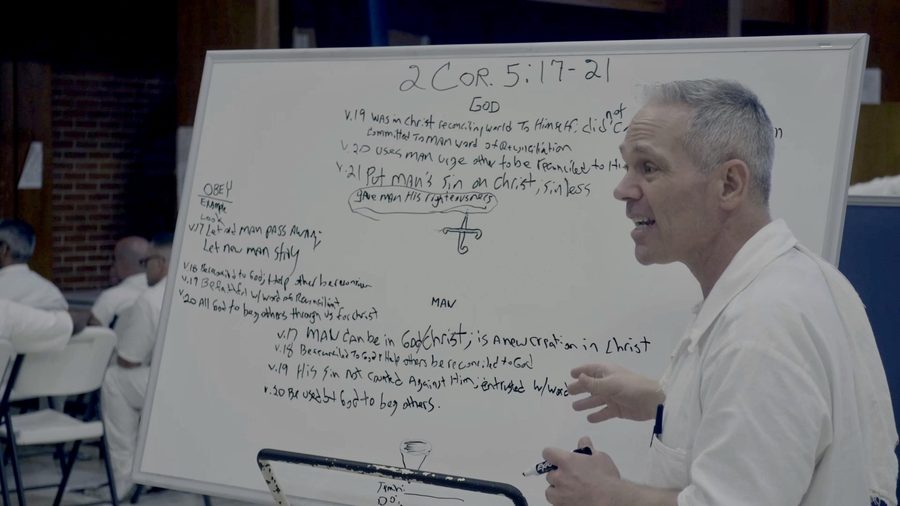 A man in prison whites teaching from a whiteboard covered in notes on 2 Corinthians 5:17-21